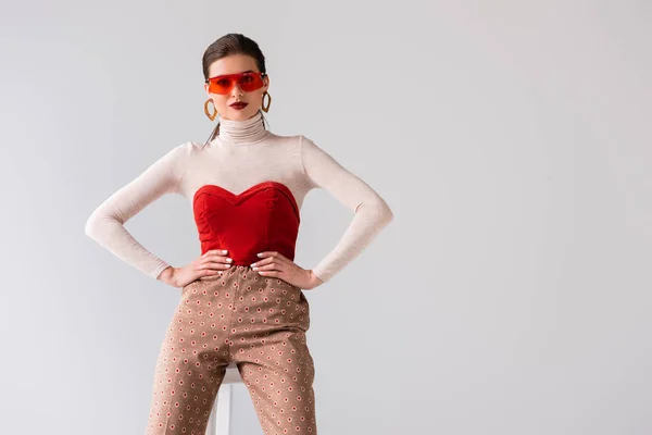 Fille à la mode tenant la main sur les hanches tout en s'appuyant sur les selles et en regardant la caméra isolée sur gris — Photo de stock