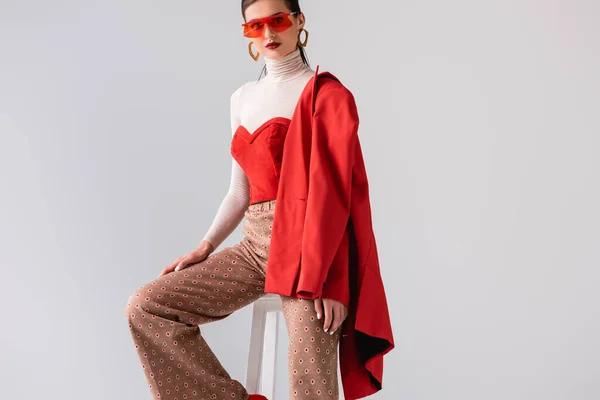 Fashionable girl with red blazer on shoulder sitting on stool and looking at camera isolated on grey — Stock Photo