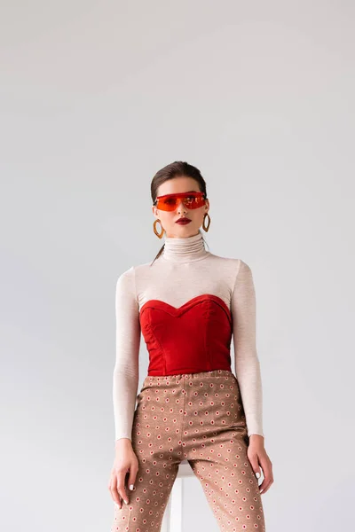 Trendy girl in sunglasses looking at camera while leaning on stool isolated on grey — Stock Photo