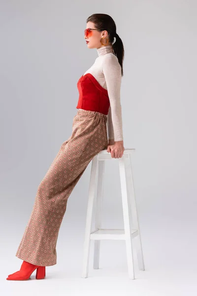 Side view of fashionable girl in beige trousers and red corset on turtleneck leaning on stool on grey — Stock Photo