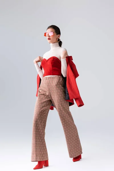 Full length view of stylish girl in sunglasses holding red blazer while walking on grey — Stock Photo