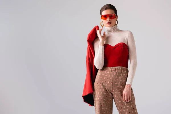 Chica de moda en gafas de sol sosteniendo chaqueta roja mientras mira a la cámara aislada en gris - foto de stock