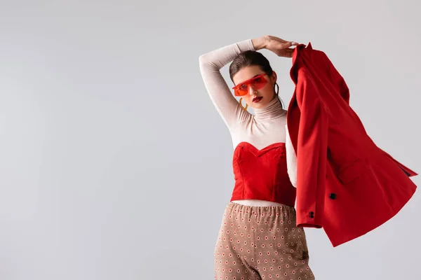 Menina na moda em óculos de sol segurando blazer vermelho enquanto olha para a câmera isolada no cinza — Fotografia de Stock
