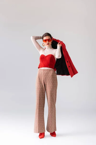 Vue pleine longueur de fille élégante dans les lunettes de soleil tenant blazer rouge sur gris — Photo de stock