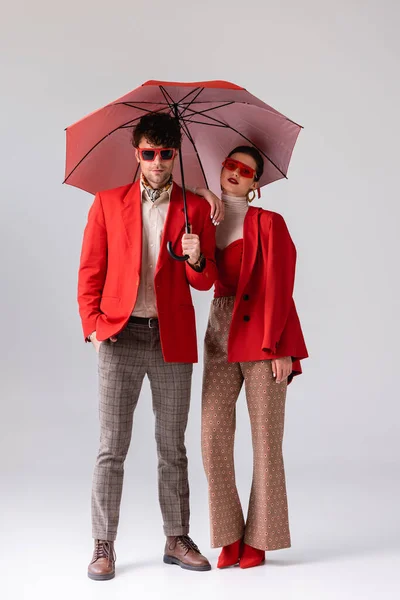 Visão comprimento total do jovem casal na moda em blazers vermelhos posando com guarda-chuva em cinza — Fotografia de Stock