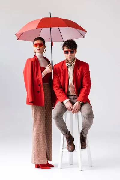 Vue pleine longueur de femme tendance tenant parapluie près de l'homme élégant assis sur tabouret sur gris — Photo de stock