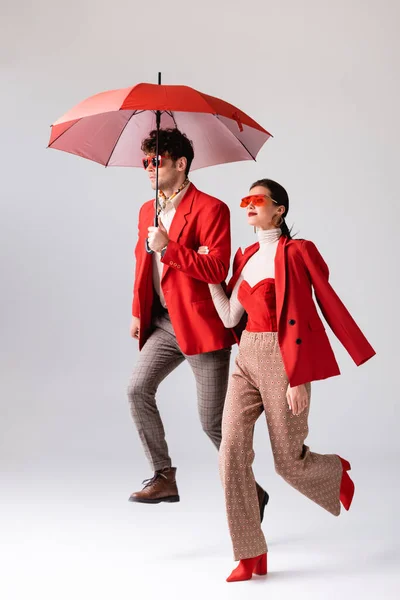 Full length view of trendy couple in red blazers and sunglasses running with umbrella on grey — Stock Photo