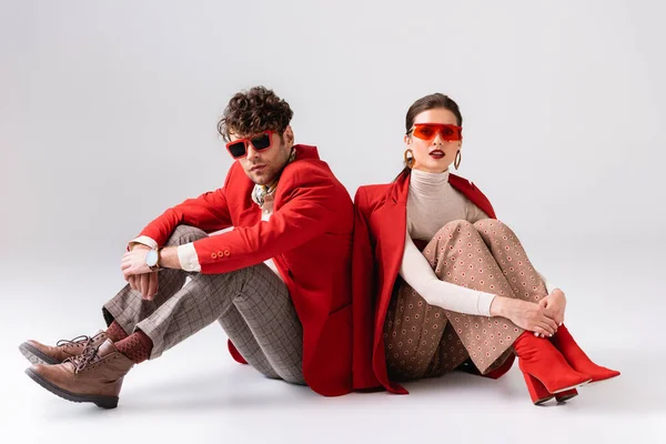 Pareja de moda en blazers rojos y gafas de sol sentados en el suelo mientras posan en gris - foto de stock