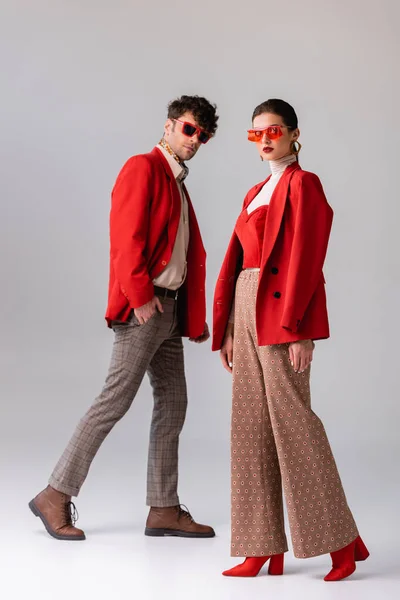 Модная пара в красных блейзерах и солнцезащитных очках в полный рост смотрит на камеру в сером — стоковое фото