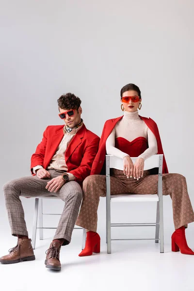 Stylish couple in sunglasses sitting on chairs and looking at camera on grey — Stock Photo