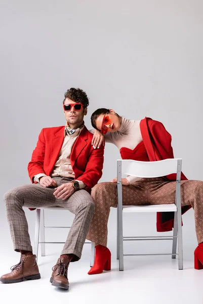 Elegant girl leaning on shoulder of trendy man while sitting on chairs on grey — Stock Photo