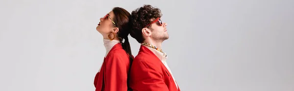 Panoramic shot of trendy couple in red blazers and sunglasses standing back to back isolated on grey — Stock Photo