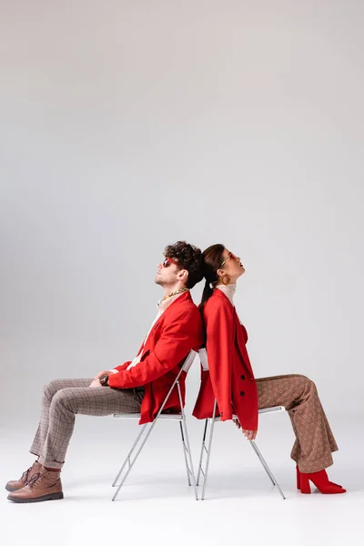 Vista lateral de pareja de moda en ropa de otoño sentado en sillas espalda con espalda en gris - foto de stock