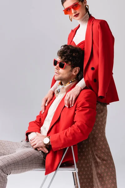 Attractive, trendy woman touching shoulders of fashionable man sitting on chair on grey — Stock Photo