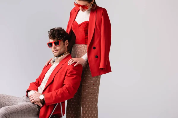 Femme élégante touchant les épaules de l'homme à la mode assis sur une chaise isolée sur gris — Photo de stock