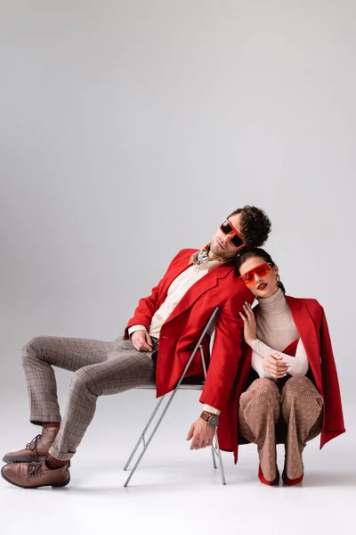 Elegante Frau hockt und berührt trendigen Mann sitzt auf Stuhl auf grau — Stockfoto