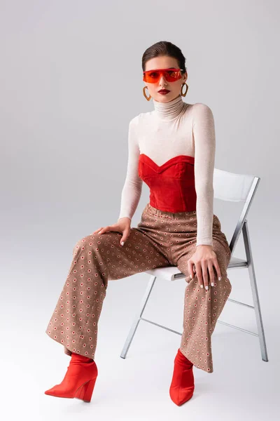 Femme élégante en lunettes de soleil rouges regardant la caméra et assis sur la chaise sur gris — Photo de stock