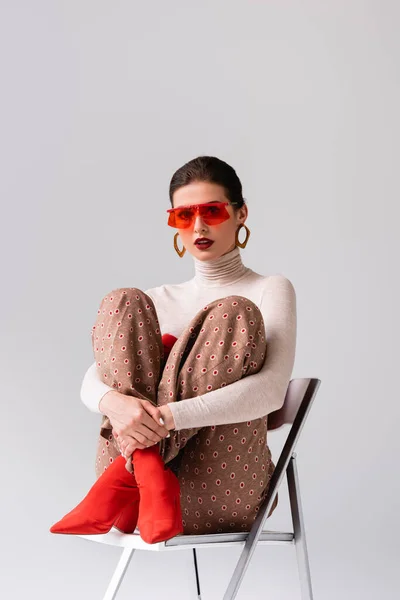 Elegant girl looking at camera and embracing crossed legs, sitting on chair isolated on grey — Stock Photo