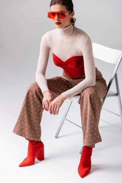 Mujer de moda inclinada hacia adelante y mirando hacia otro lado, sentada en la silla en gris - foto de stock