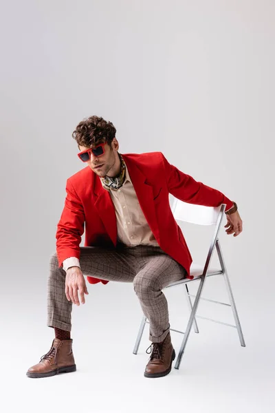 Стильний чоловік у червоному блістері спирається вперед і позує, сидячи на стільці на сірому — стокове фото