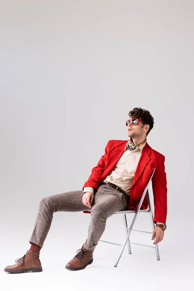 Elegante uomo in blazer rosso appoggiato indietro e in posa mentre seduto sulla sedia su grigio — Foto stock
