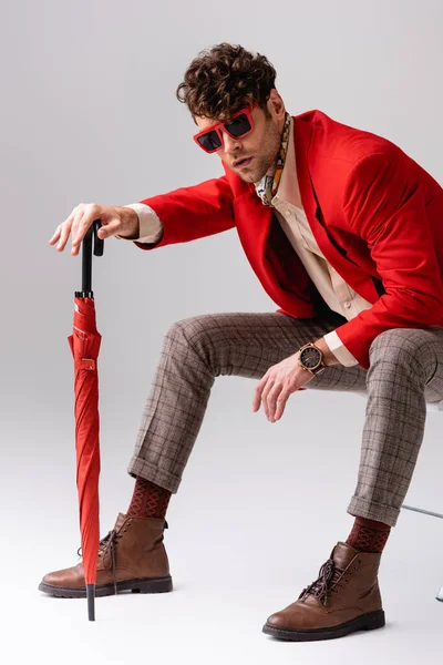 Homem na moda em blazer vermelho inclinado para a frente e posando com guarda-chuva enquanto sentado na cadeira em cinza — Fotografia de Stock