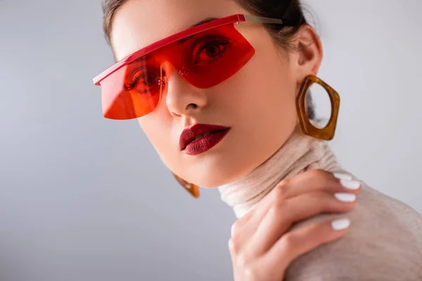 Attraente ragazza in occhiali da sole rossi toccare spalla e guardando la fotocamera isolata su grigio — Stock Photo