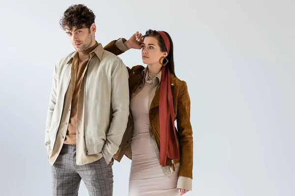 Trendiges Mädchen im Herbst-Outfit posiert in der Nähe von stilvollen Mann mit Händen in der Tasche isoliert auf grau — Stockfoto