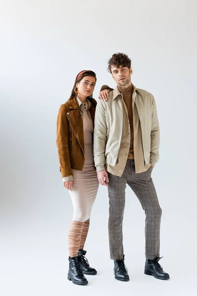 Vista completa de la mujer con estilo en traje de otoño posando cerca del hombre de pie con la mano en el bolsillo en gris - foto de stock