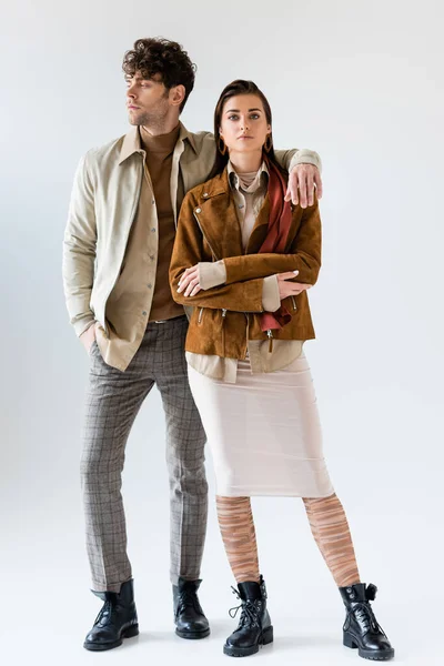 Полный обзор стильного мужчины с рукой в кармане, глядя в сторону, обнимая привлекательную женщину, стоящую рядом на сером — стоковое фото