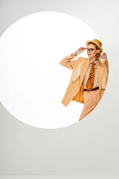 Stylish woman in straw hat and beige jacket adjusting eyeglasses near circle on white background — Stock Photo