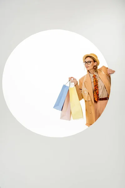 Mujer elegante sonriente sosteniendo bolsas de compras detrás de agujero redondo sobre fondo blanco - foto de stock