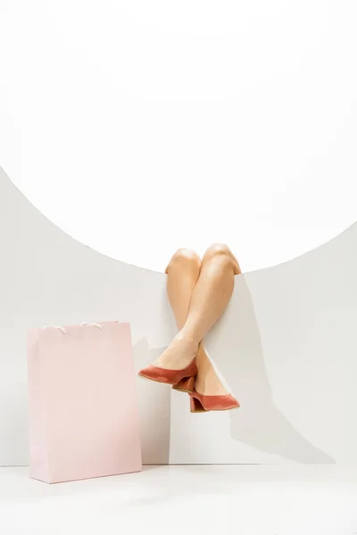 Cropped view of female legs in coral shoes in round hole near shopping bag on white background — Stock Photo