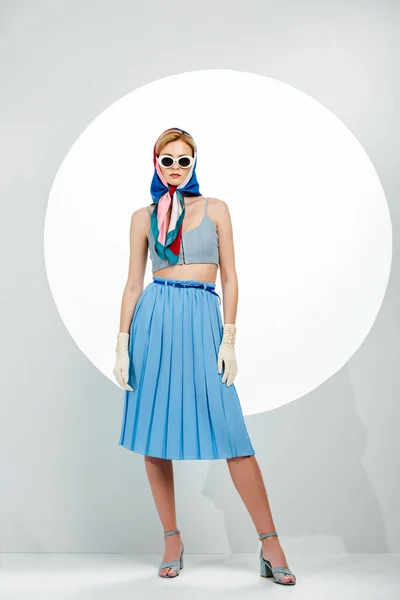Mulher elegante em óculos de sol e saia azul em pé perto do círculo no fundo branco — Fotografia de Stock