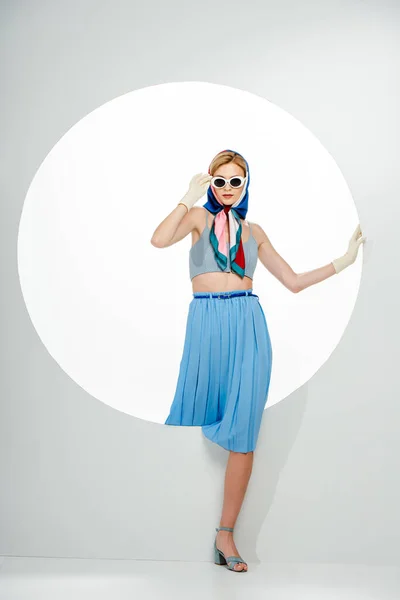 Mulher na moda em lenço de cabeça ajustando óculos e tocando buraco redondo no fundo branco — Fotografia de Stock