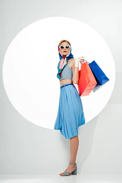 Mulher na moda em óculos de sol e lenço de cabeça segurando sacos de compras azuis e vermelhos perto do círculo no fundo branco — Fotografia de Stock