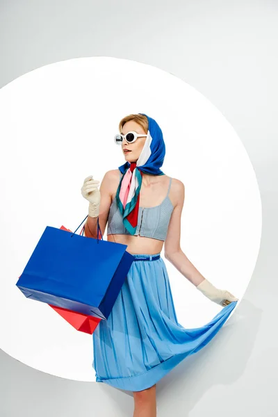 Модная девушка в перчатках и солнцезащитных очках с синими и красными сумками для покупок на белом фоне — Stock Photo