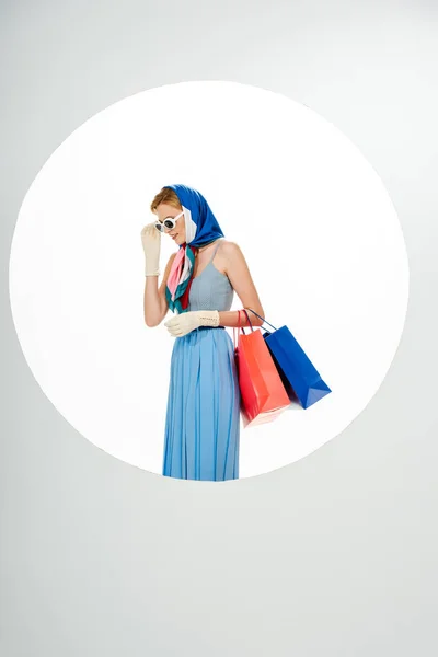 Mujer elegante sonriendo mientras se ajustan las gafas de sol y la celebración de coloridas bolsas de compras cerca del círculo sobre fondo blanco - foto de stock