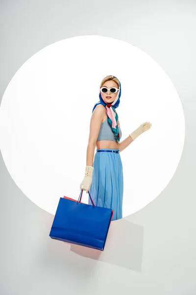 Chica de moda en pañuelo para la cabeza y gafas de sol sosteniendo bolsas de compras cerca del círculo sobre fondo blanco - foto de stock