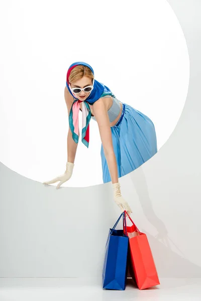 Trendy woman in headscarf and sunglasses putting red and blue shopping bags near circle on white background — Stock Photo