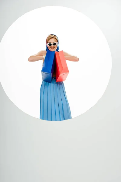 Mulher alegre em óculos de sol e lenço de cabeça segurando sacos de compras vermelhos e azuis perto do círculo no fundo branco — Fotografia de Stock