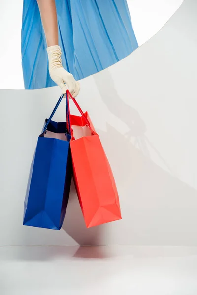 Vue recadrée d'une femme élégante dans un gant tenant des sacs à provisions rouges et bleus près d'un trou rond sur fond blanc — Photo de stock