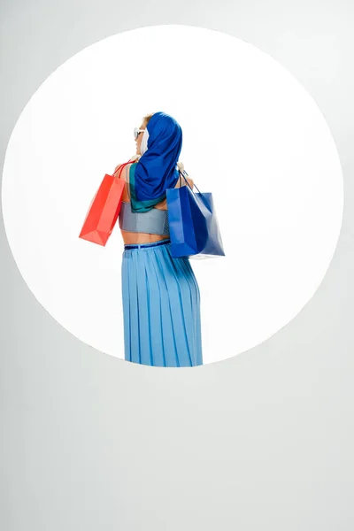 Vista posterior de la mujer con estilo en pañuelo para la cabeza con bolsas de compras rojas y azules cerca del círculo sobre fondo blanco - foto de stock