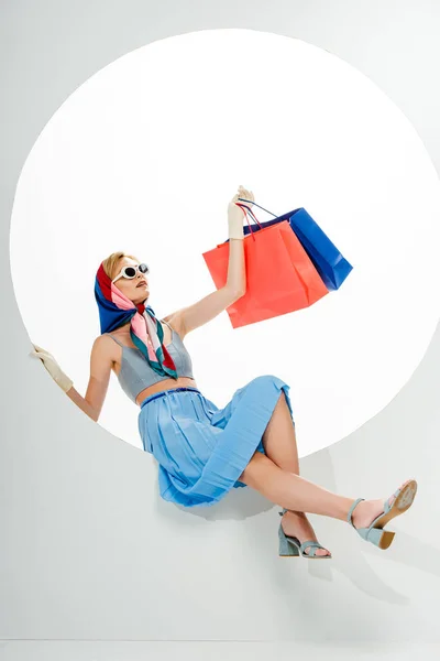 Mujer de moda en gafas de sol posando mientras que las bolsas de compras azules y rojas en círculo sobre fondo blanco - foto de stock