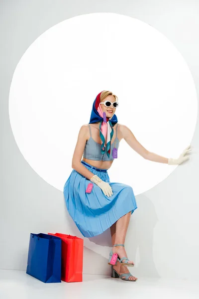 Mujer elegante sonriente en gafas de sol con etiquetas de precio en la ropa sentada cerca de bolsas de compras en círculo sobre fondo blanco - foto de stock