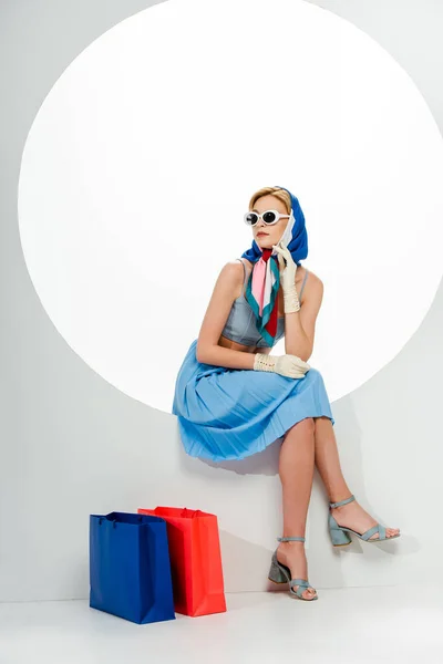 Fille à la mode en lunettes de soleil et foulard assis en cercle près de sacs à provisions sur fond blanc — Photo de stock