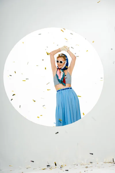 Femme souriante en foulard et lunettes de soleil debout sous les confettis tombant près du trou rond sur fond blanc — Photo de stock