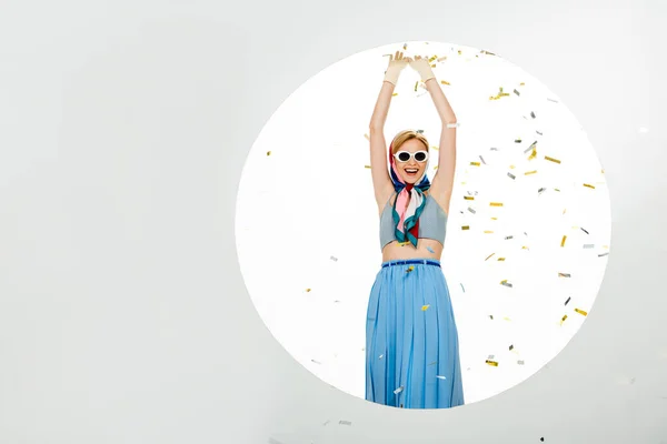 Позитивная девушка в солнечных очках, стоящая возле круга под падающими конфетти на белом фоне — стоковое фото