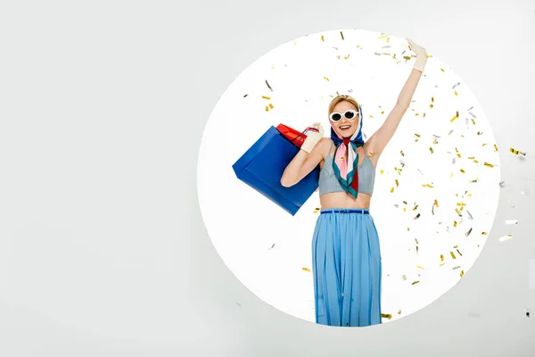 Усміхнена жінка в сонцезахисних окулярах тримає барвисті сумки для покупок під падінням конфетті біля кола на білому тлі — стокове фото