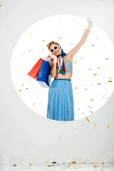Усміхнена жінка в окулярах і сонцезахисних окулярах тримає сумки під падінням конфетті біля кола на білому тлі — стокове фото